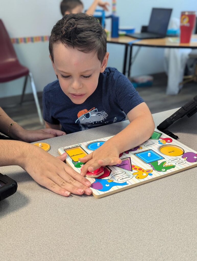 A young boy sits at a table, focusing on placing puzzle pieces on a colorful educational board, guided by an adult’s hand.