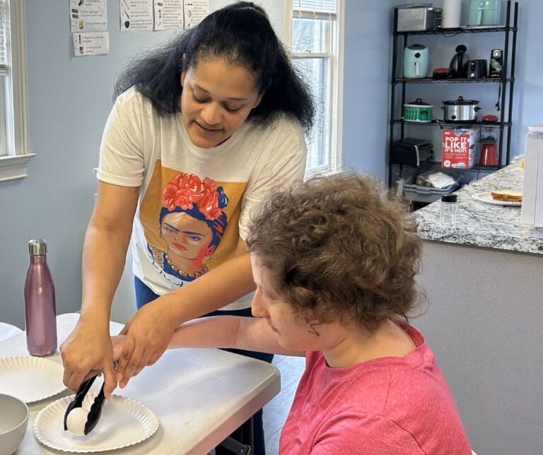 A woman helps another person use tongs to place food on a paper plate in a kitchen setting.