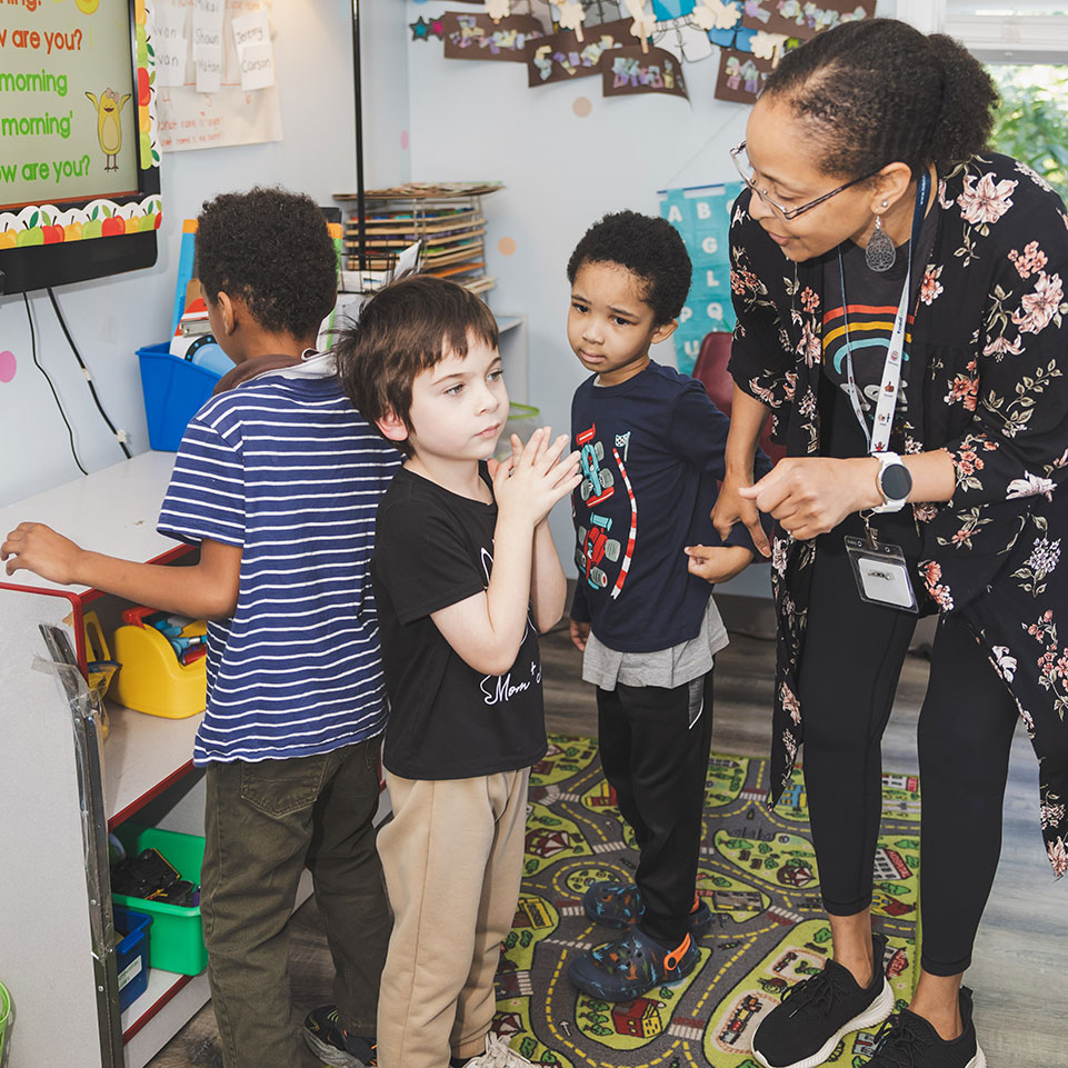 A teacher interacts with three young children standing in line in a colorful classroom, with educational materials and a screen visible in the background.