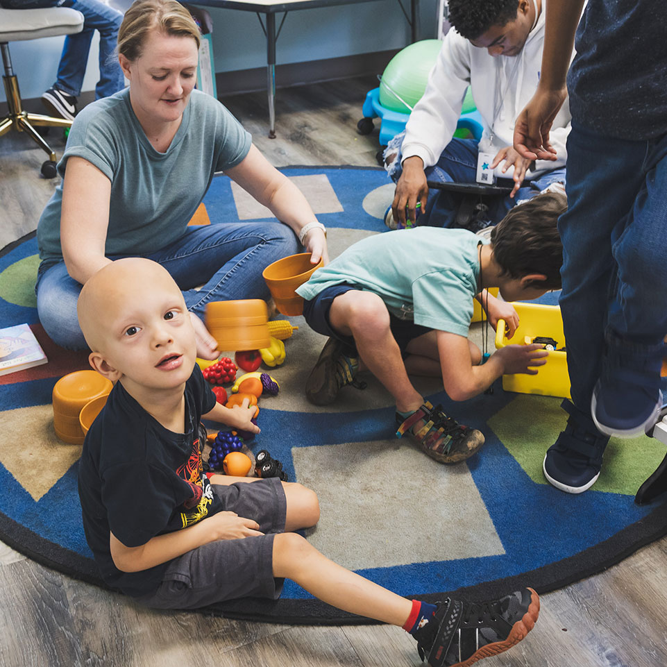A group of children play with toys on a colorful rug, supervised by adults in a classroom setting. One child sits and looks toward the camera.