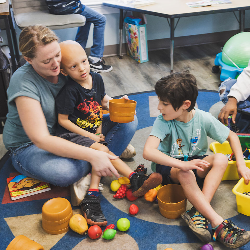 An adult and two children sit on a classroom rug, playing with plastic fruits and toy bins. The adult points while one child holds a container.