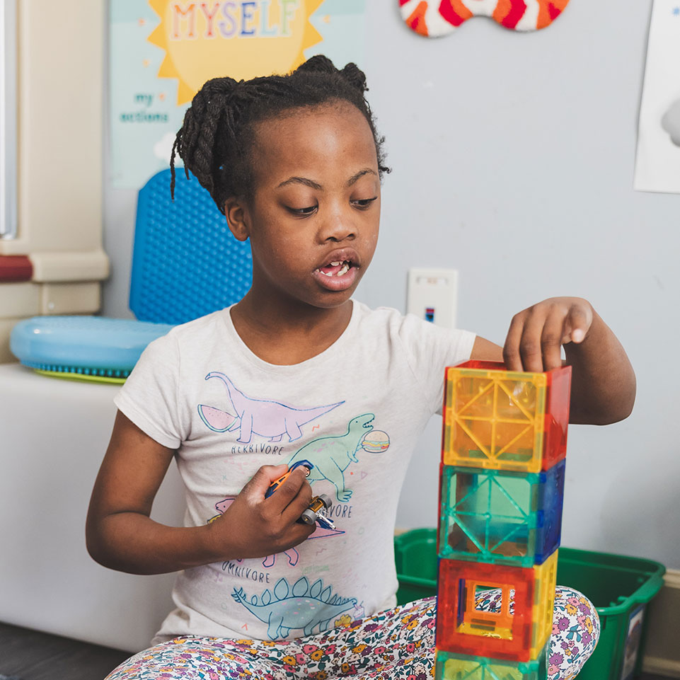 A young girl builds a tall structure with colorful magnetic tiles while sitting on the floor in a classroom or playroom.