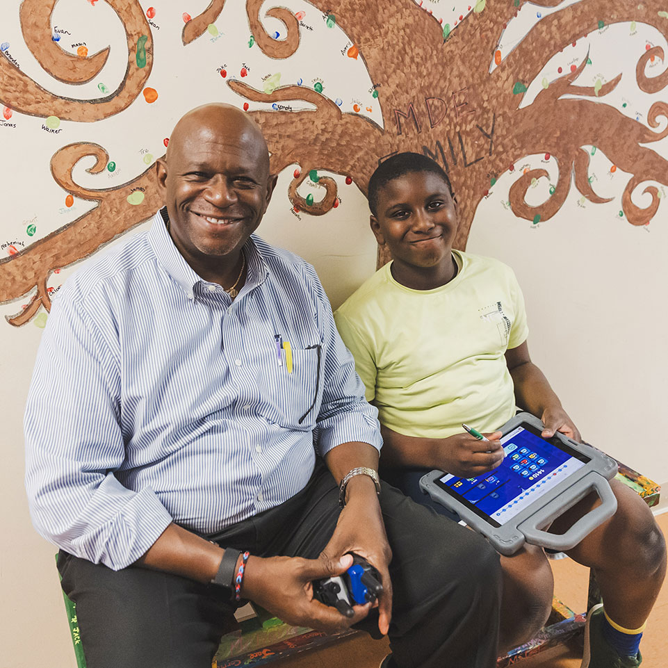 An adult and a child sit on a bench in front of a mural. The child holds a tablet and stylus while both smile at the camera.