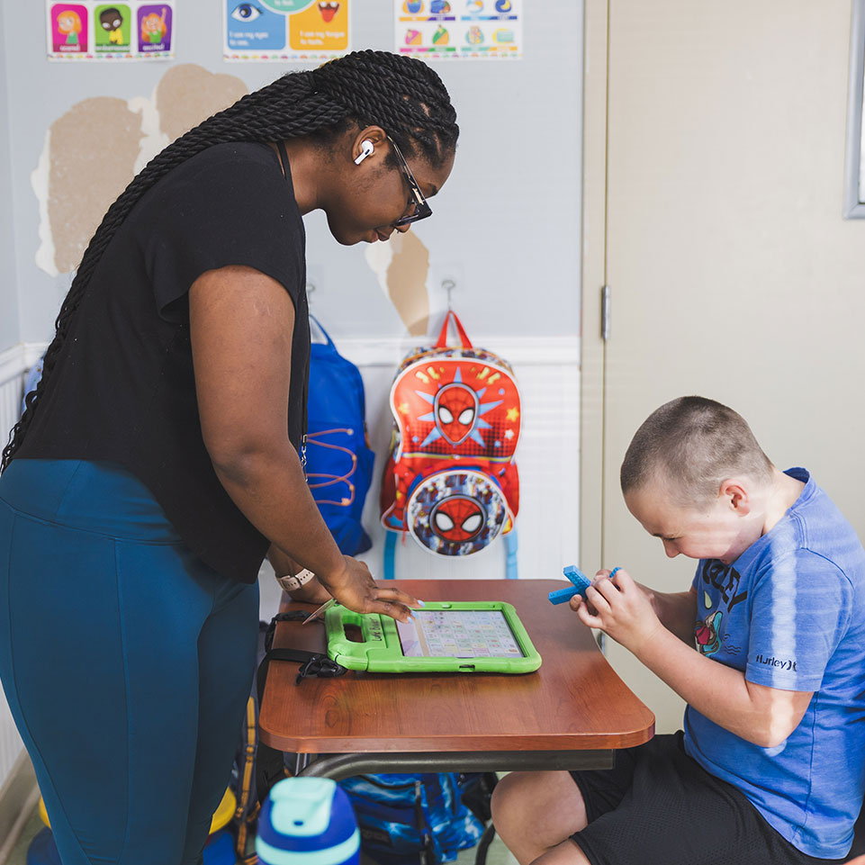 An adult helps a child use a tablet at a small table in a classroom; colorful backpacks and posters are visible in the background.