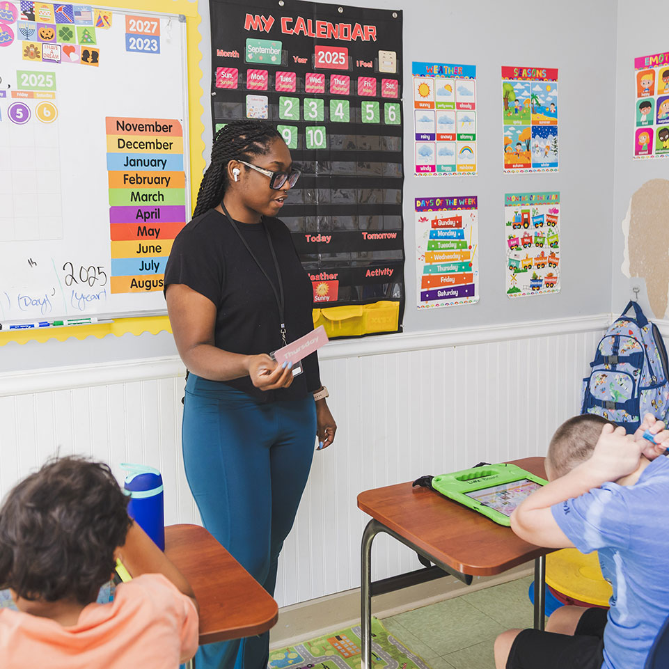A teacher stands at the front of a classroom holding a card while two young students sit at desks, surrounded by educational posters and a calendar on the wall.