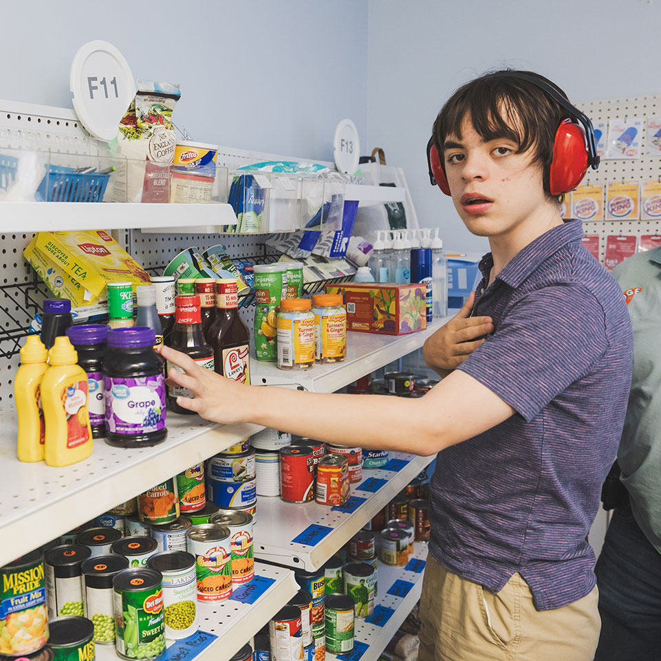A person wearing red ear protectors selects items from shelves stocked with canned goods, condiments, and packaged foods in a store or pantry setting.