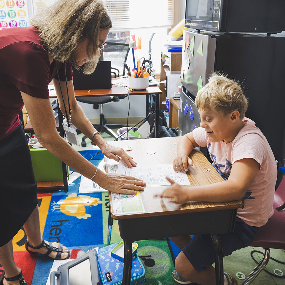 A teacher leans over a desk to assist a smiling student with schoolwork in a classroom filled with supplies and colorful decorations.