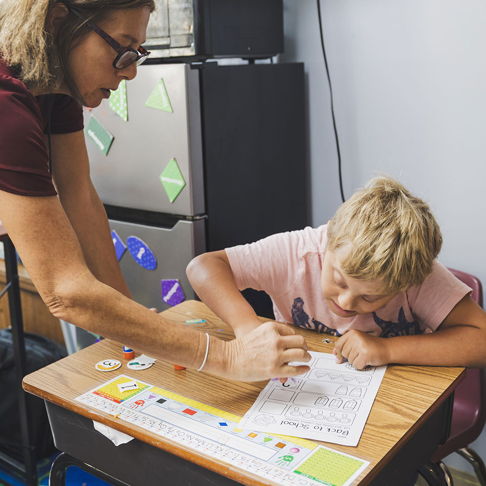 An adult helps a young boy with a worksheet at a classroom desk. The boy is focused on the paper while the adult points to something on the sheet.