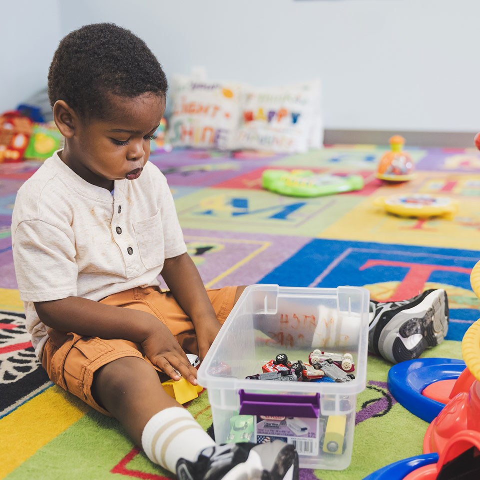 Young boy sits on a colorful alphabet rug, playing with toy cars from a plastic container in a playroom.