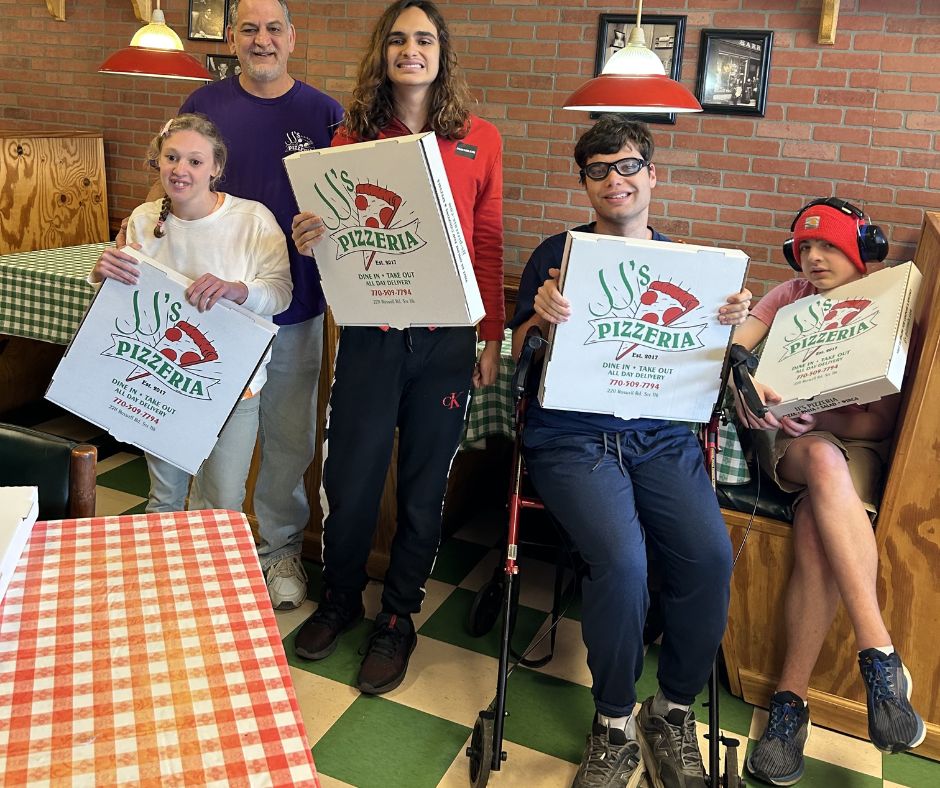 Five people stand and sit inside a pizzeria, holding large pizza boxes from JJ’s Pizzeria, with checkered tablecloths and wood-paneled booths in the background.