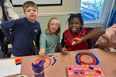 Three children sit at a table painting colorful circles on paper, smiling at the camera. Paints and brushes are scattered on the table.