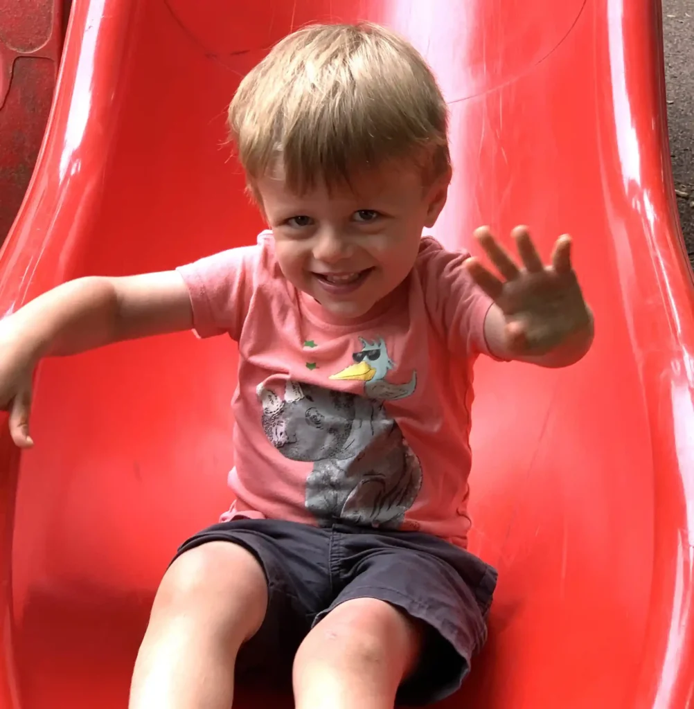 Young child with light brown hair, wearing a pink animal-print shirt and dark shorts, sits on a red playground slide, smiling and waving at the camera.