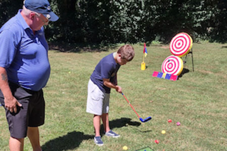 An adult watches a child using a golf club to hit balls toward targets on a grassy field, with colorful targets and equipment in the background.