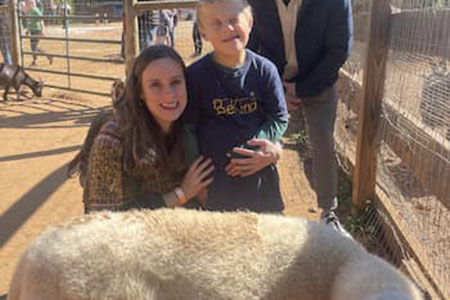 A woman and a boy stand next to a large white animal at a petting zoo, with a man standing behind them and goats visible in the background.
