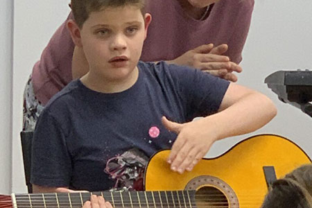 A boy in a dark t-shirt gestures with his hand above the strings of a yellow acoustic guitar while another person stands behind him.