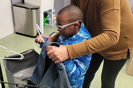 A child wearing glasses and a blue jacket holds a denim jacket and hanger, assisted by an adult in a brown sweater.