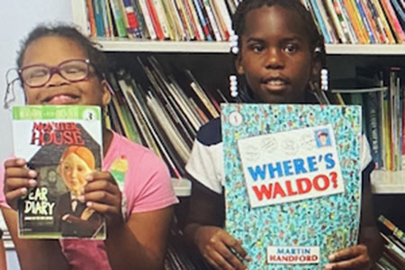 Two children stand in front of bookshelves, each holding a book. The child on the left holds "Car Diary," and the child on the right holds "Where's Waldo?.