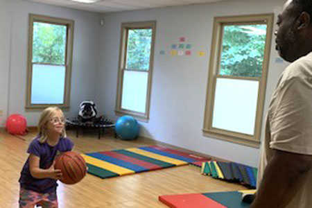A young girl holding a basketball prepares to make a pass to an adult in a room with gym mats and exercise balls.