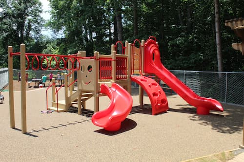 Outdoor playground with red slides, climbing structures, monkey bars, and a bridge, set on a safety surface and surrounded by trees and a chain-link fence.