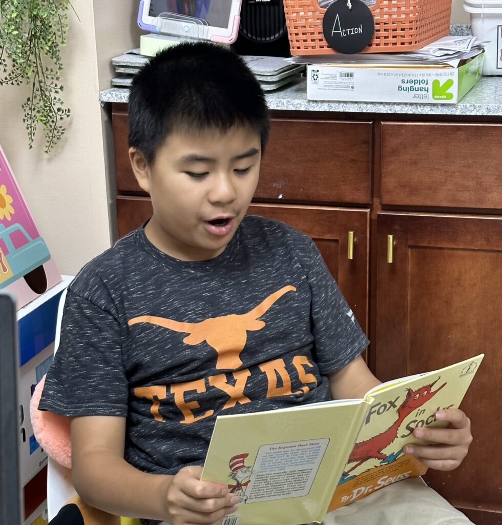 A boy sits indoors reading "Fox in Socks" by Dr. Seuss aloud. He is wearing a dark T-shirt with "TEXAS" and a longhorn logo on it.