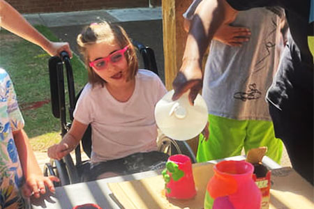 A child in a wheelchair wearing pink glasses sits at a table outdoors while another person pours liquid into a colorful container.