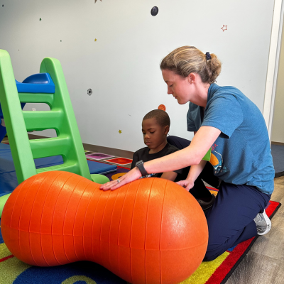 An adult helps a child balance on an orange peanut-shaped exercise ball in a playroom with a green slide and colorful mats.