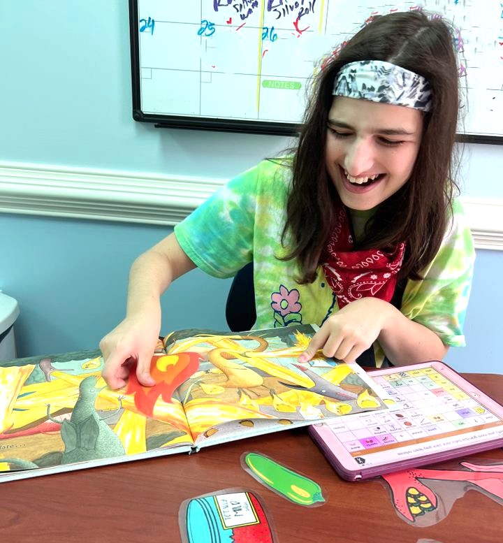 A person wearing a tie-dye shirt and bandana smiles while pointing at a picture book, with an augmentative communication tablet on the table.