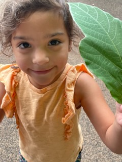 Young girl in an orange sleeveless shirt smiles at the camera while holding a large green leaf outdoors.