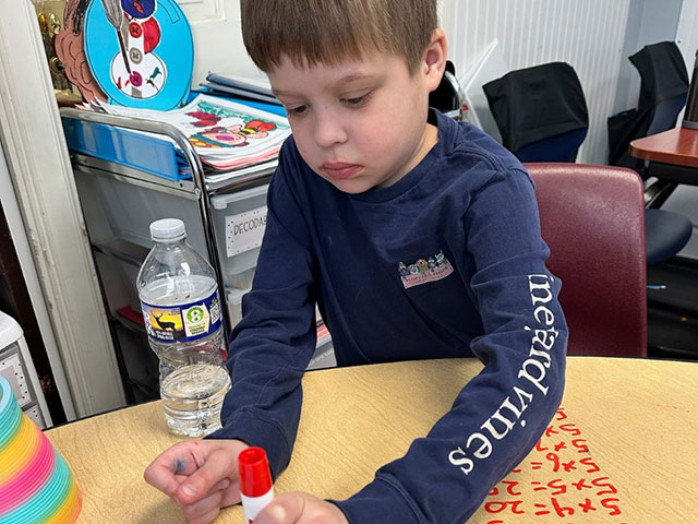 A boy sits at a table holding a red marker, with a water bottle and a colorful activity sheet nearby.