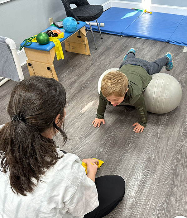 A child lies face down on an exercise ball with their hands on the floor while an adult observes in a therapy room with equipment in the background.