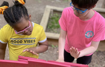Two children stand at an outdoor play structure, both wearing glasses. One wears a yellow "Good Vibes" shirt, the other a pink shirt. They play on a red musical panel.
